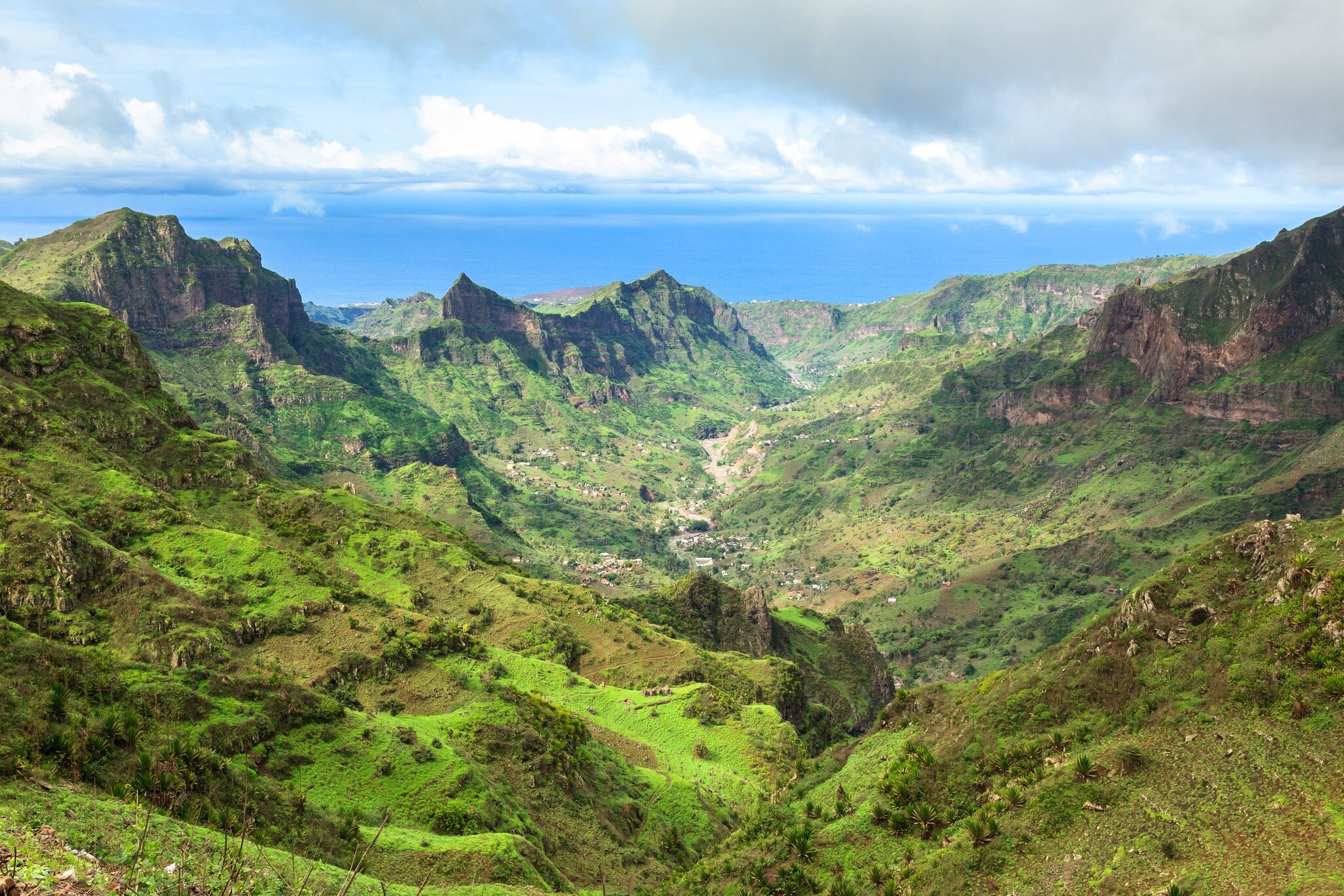 Steep green mountain ridges and a deep valley dotted with houses, the Atlantic Ocean on the horizon under clouds