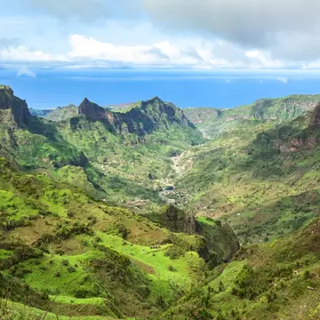 Steep green mountain ridges and a deep valley dotted with houses, the Atlantic Ocean on the horizon under clouds