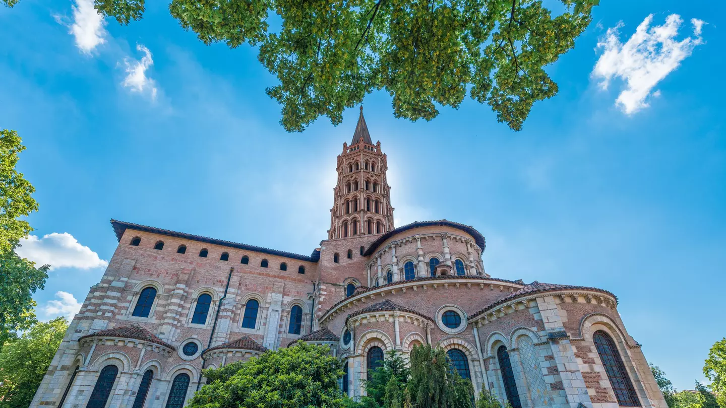 View of the Basilica of St-Sernin, where a pink and white church with an ornate steeple is backlit by the sun on a blue-sky day