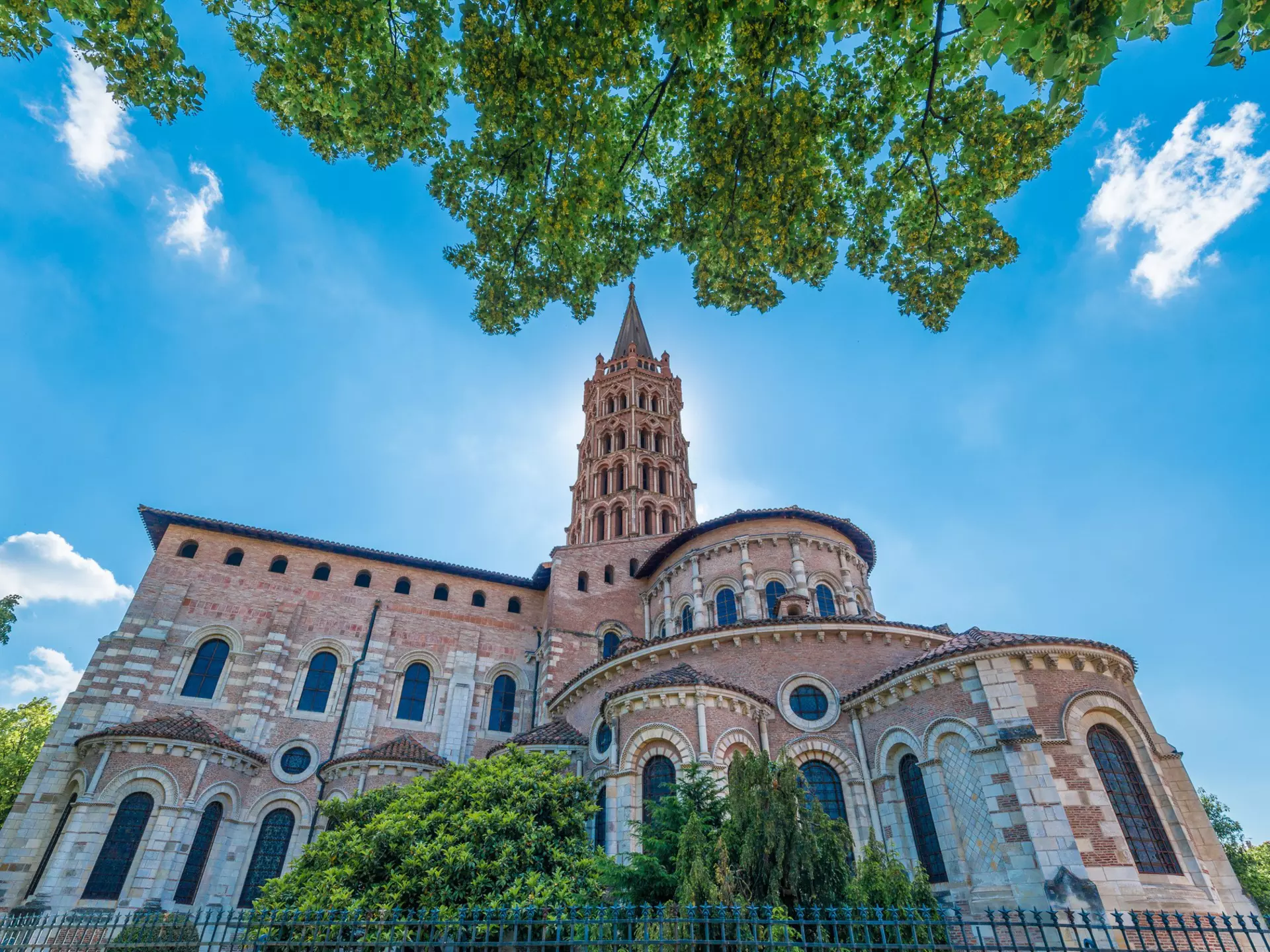 View of the Basilica of St-Sernin, where a pink and white church with an ornate steeple is backlit by the sun on a blue-sky day