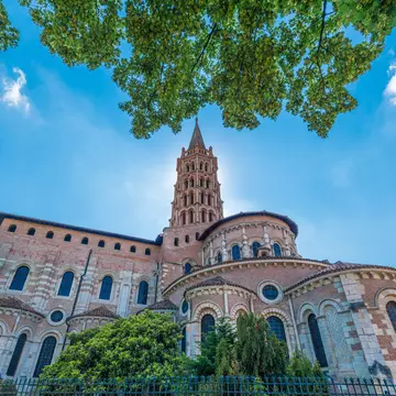 View of the Basilica of St-Sernin, where a pink and white church with an ornate steeple is backlit by the sun on a blue-sky day