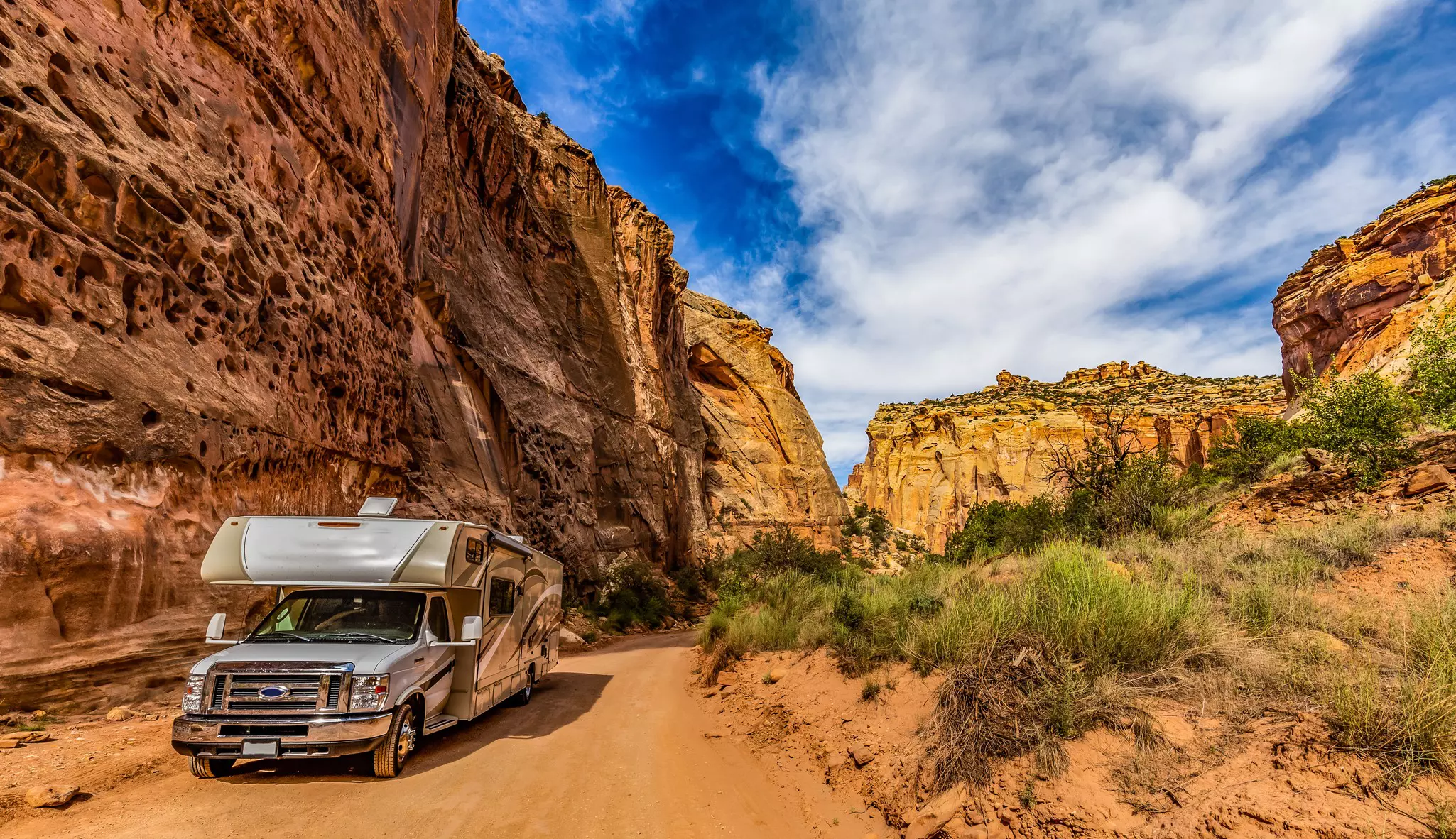 A camper van on a dusty trail along a sheer yellow and red rock face.