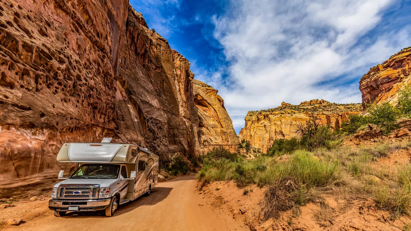 A camper van on a dusty trail along a sheer yellow and red rock face.