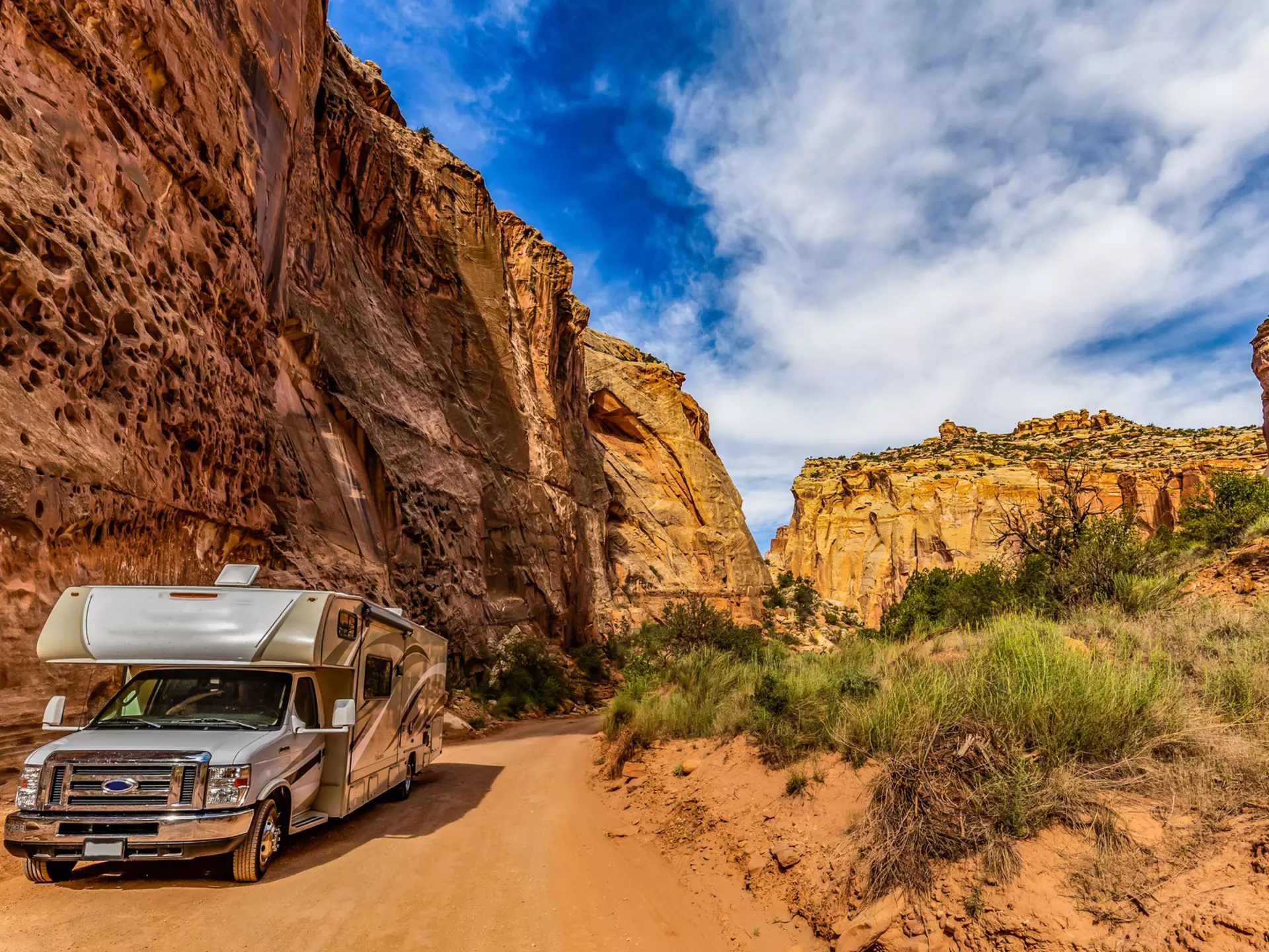 A camper van on a dusty trail along a sheer yellow and red rock face.
