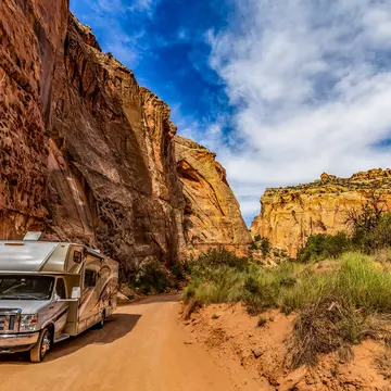 A camper van on a dusty trail along a sheer yellow and red rock face.