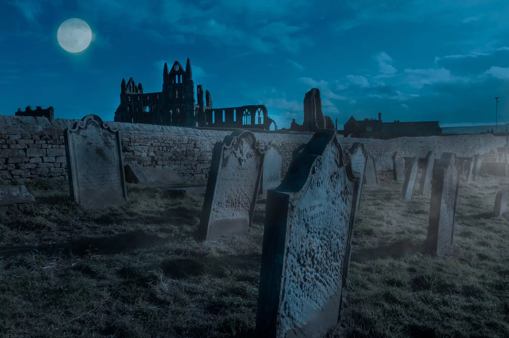 An atmospheric night-time shot of a ruined abbey with gravestones under a full moon