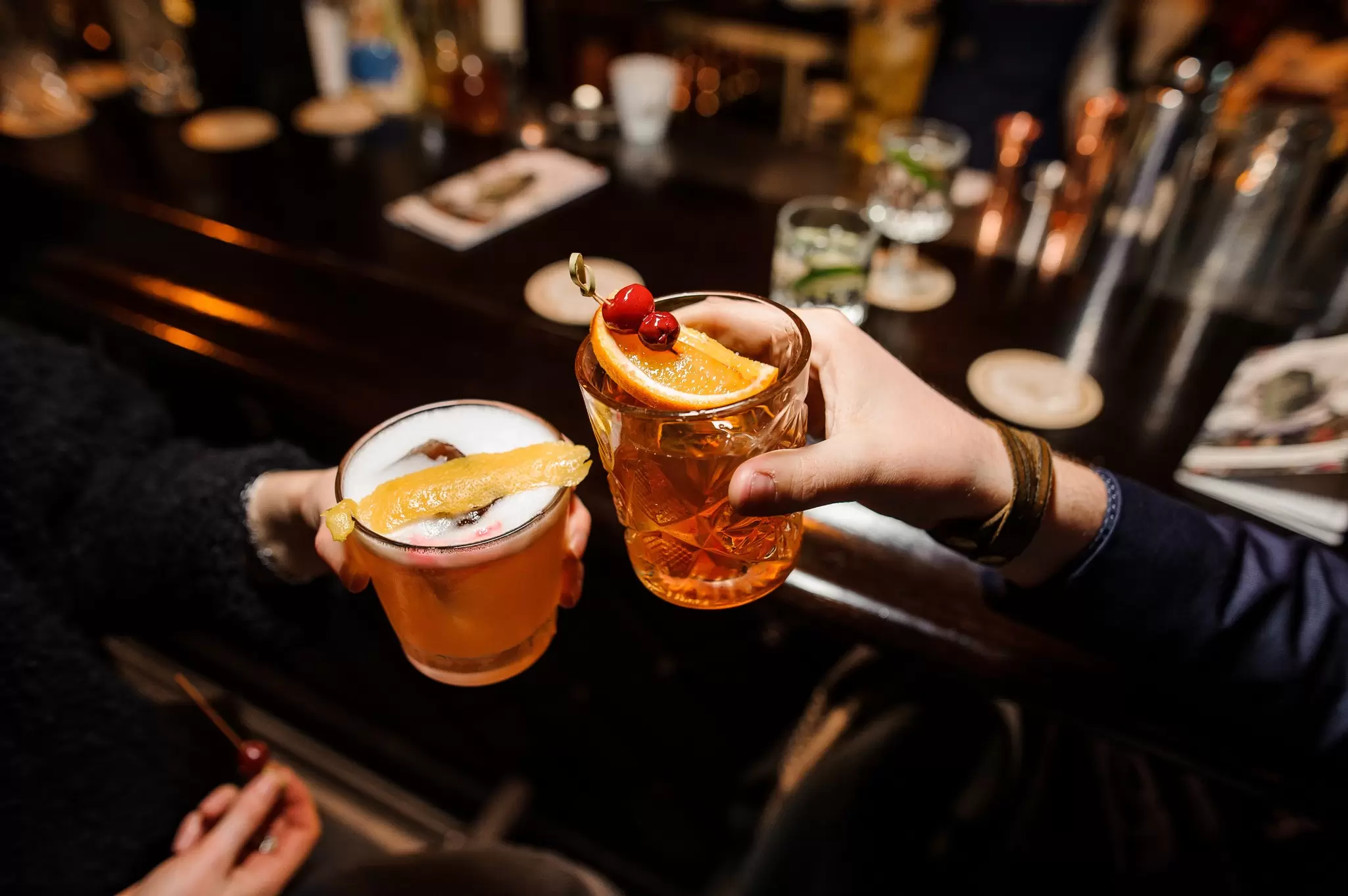 Two people clinking garnished cocktails together across a dimly lit wooden bar