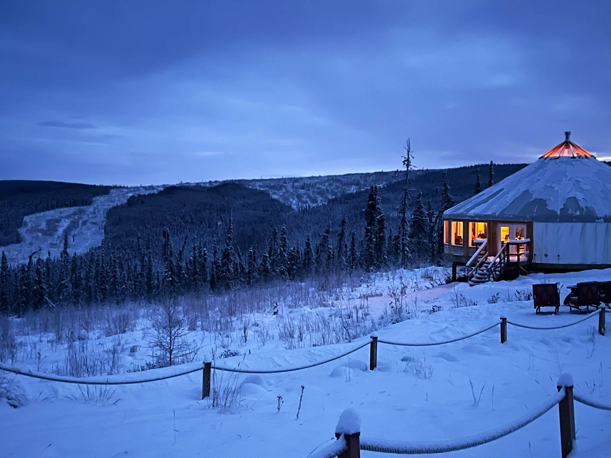 A large yurt lit from within in a snowy landscape in Alaska; two rope fences are in the foreground, and an evergreen forest is in the background.