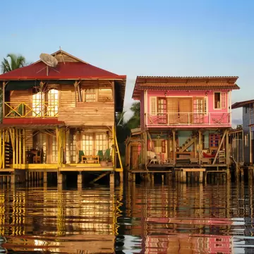 Two-storey wooden houses, one painted pink, standing on stilts over calm water with reflections, Bocas del Toro, Panama.