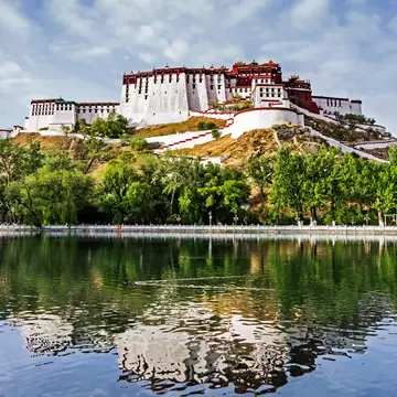 The white and red Potala Palace on its hill in Lhasa, Tibet, reflected in a still lake below a cloudy blue sky.