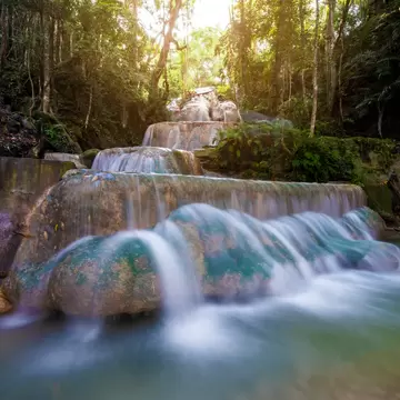 Tiered waterfall spilling over mossy limestone ledges into a pale green pool, with sunlight filtering through tall trees.