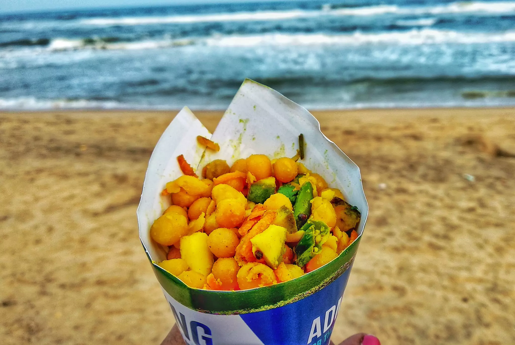 A snack of vegetables and fruit served in a paper cone and held up on a beach