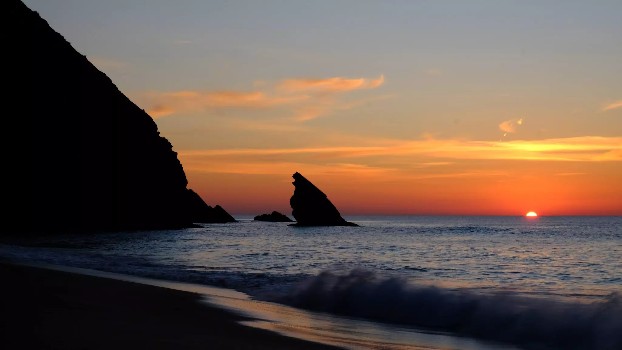 Orange sunset over the ocean with rocky hills to the left and a seastack in the distance.