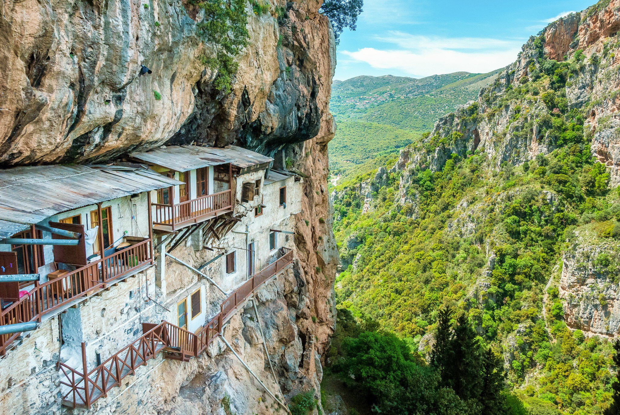 A historic monastery with balconies built into the edge of a cliff overlooking a lush valley in Greece.