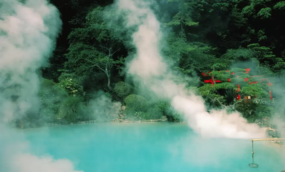 Steam rising from the turquoise Umi Jigoku hot spring pool in Beppu, red shrine buildings among trees