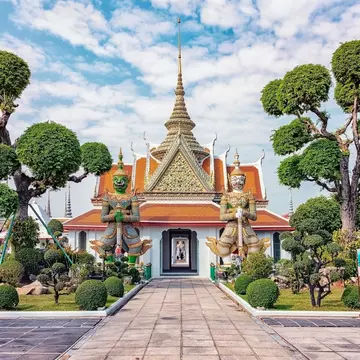 Guardian statues at Wat Arun in Bangkok, Thailand, flanked by rounded trees.