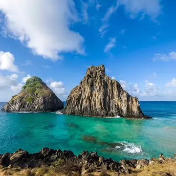 Two steep rocky islets in clear turquoise sea under a blue sky with white clouds, dark volcanic rocks and grass in front.