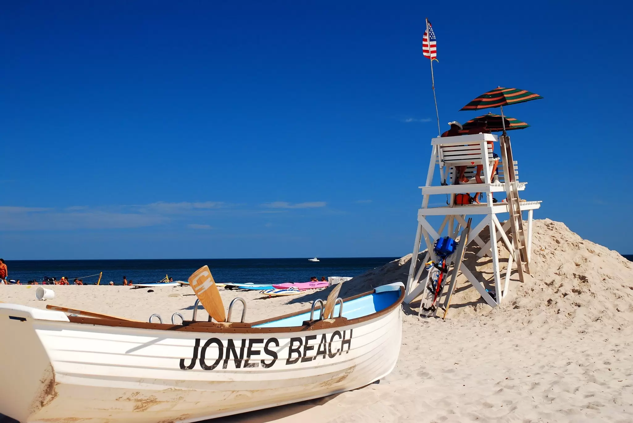 A boat painted with "Jones Beach" on its side is on the sand by a lifeguard tower.