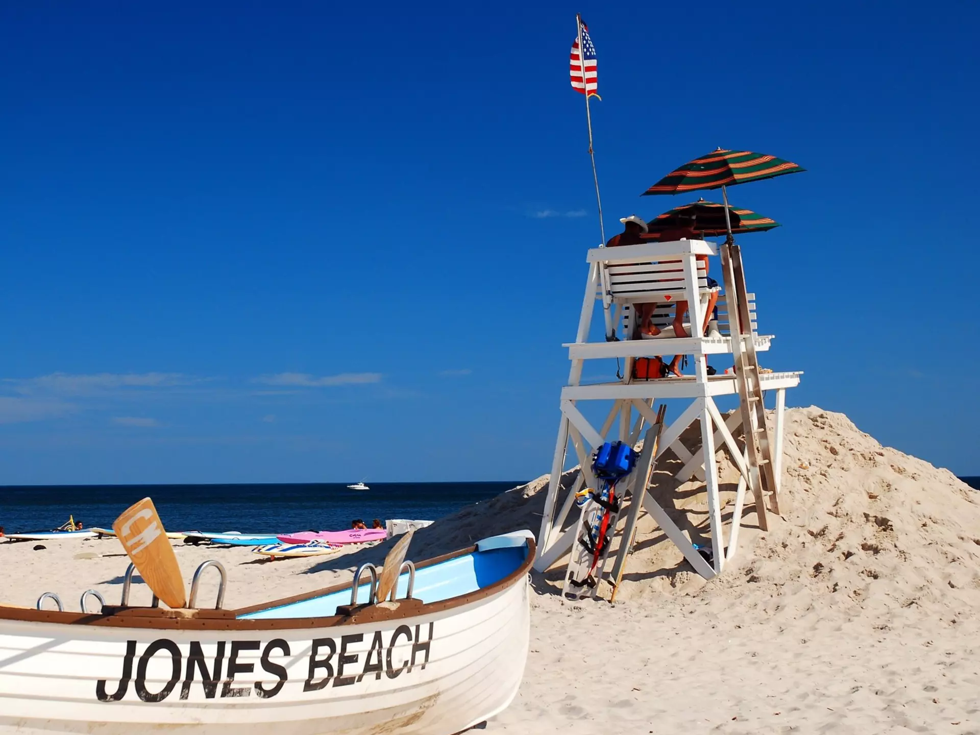 A boat painted with "Jones Beach" on its side is on the sand by a lifeguard tower.