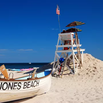 A boat painted with "Jones Beach" on its side is on the sand by a lifeguard tower.