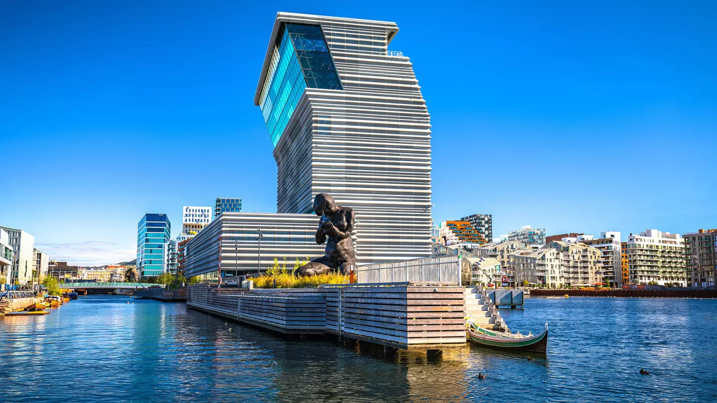 A large figurative bronze sculpture of a kneeling woman with her head bowed on a harborfront near a contemporary museum building.