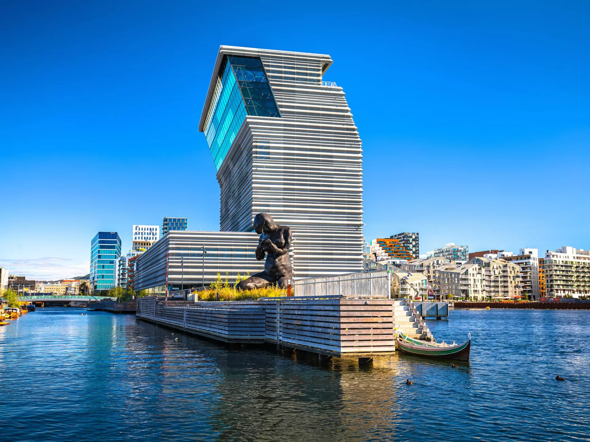 A large figurative bronze sculpture of a kneeling woman with her head bowed on a harborfront near a contemporary museum building.