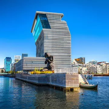 A large figurative bronze sculpture of a kneeling woman with her head bowed on a harborfront near a contemporary museum building.