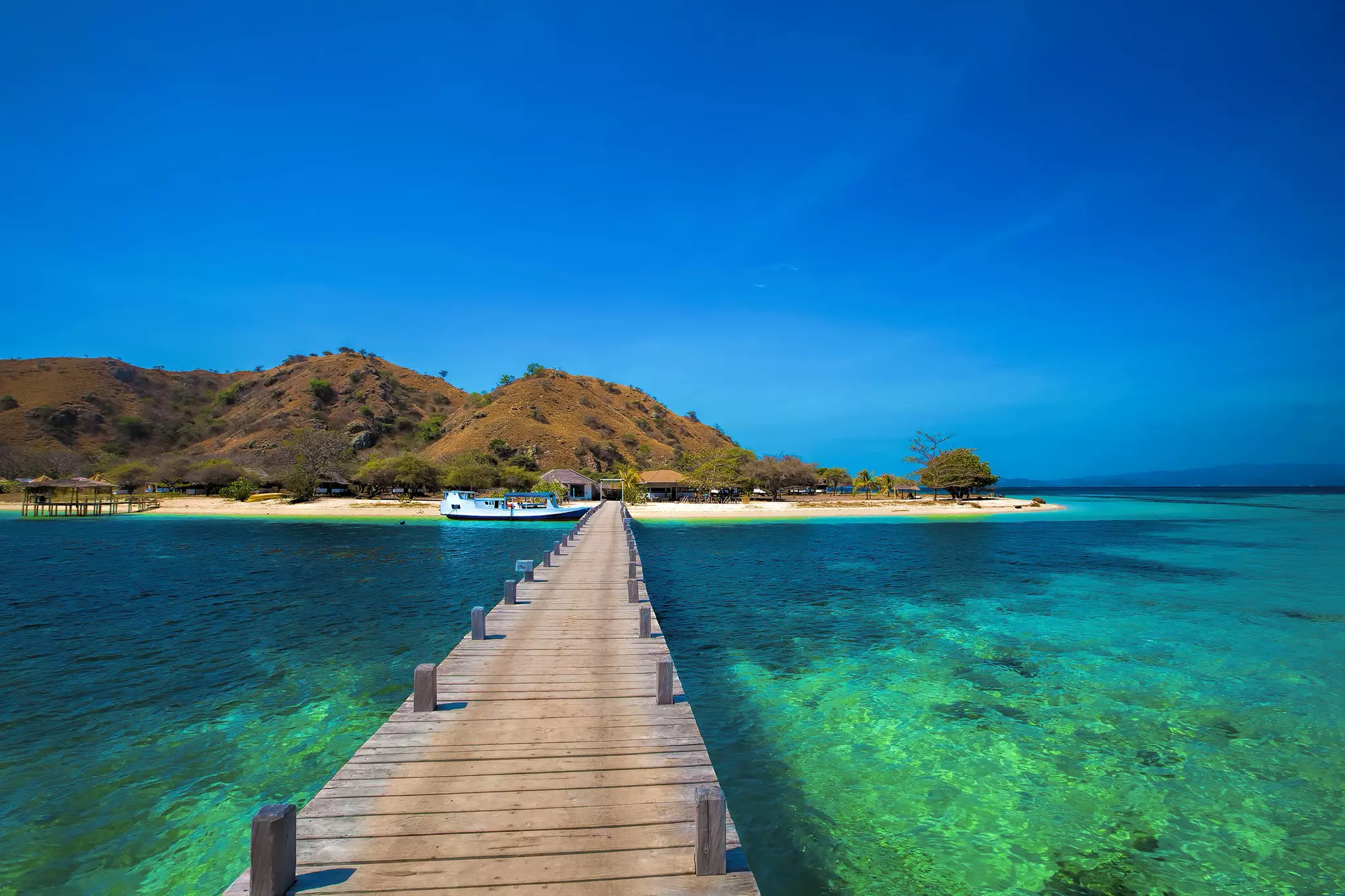 A long wooden boat jetty through green seas leading towards a sandy beach on a small island.