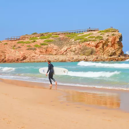 Young male surfer in dark neoprene walking on a sandy beach with white surf board. 