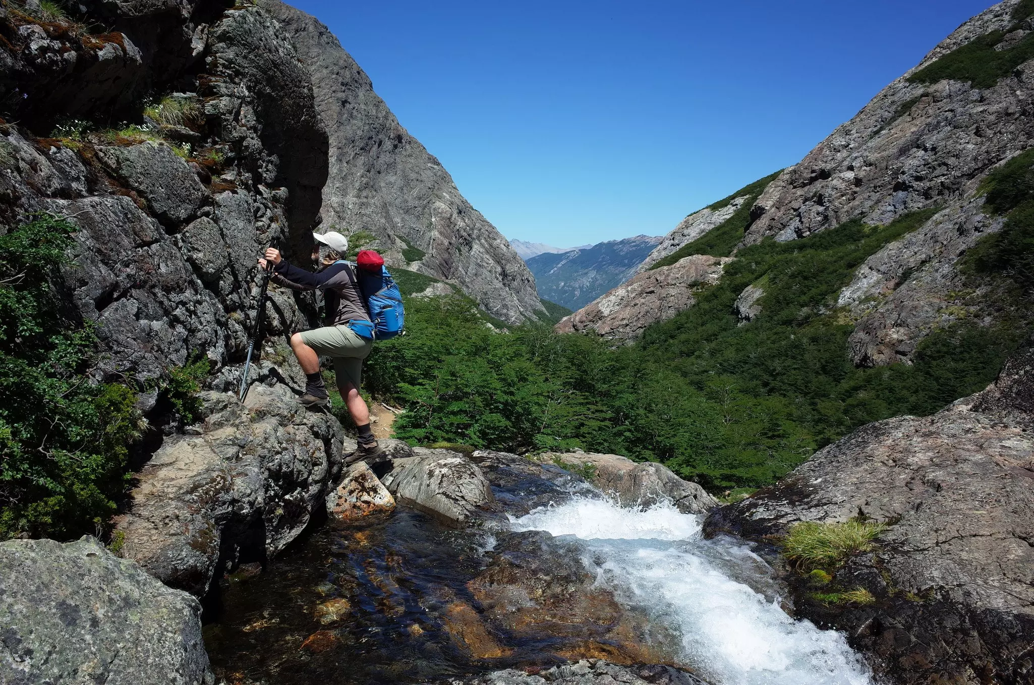 A hiker ascends on rock ledge above white water stream.