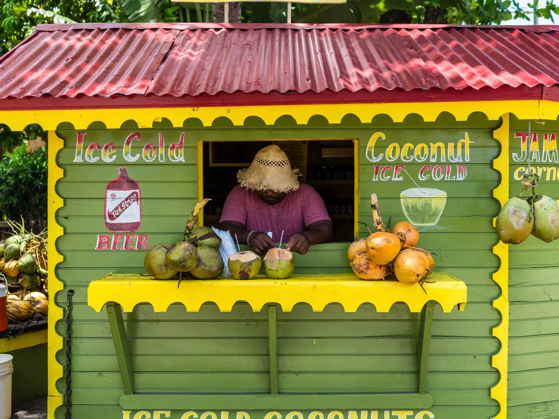 A man wearing a straw hat appears at a window in a green stand advertising beer and coconut drinks; there are coconuts on a yellow shelf in front of the window.