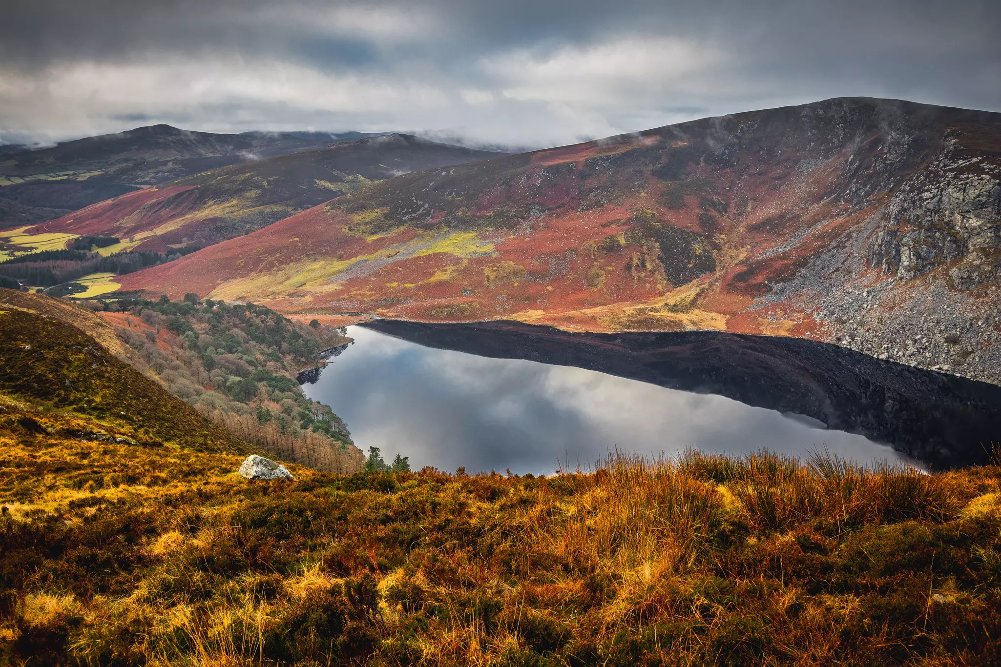 A dark lake surrounded by low hills with fall foliage of yellows and deep orange on an overcast day.
