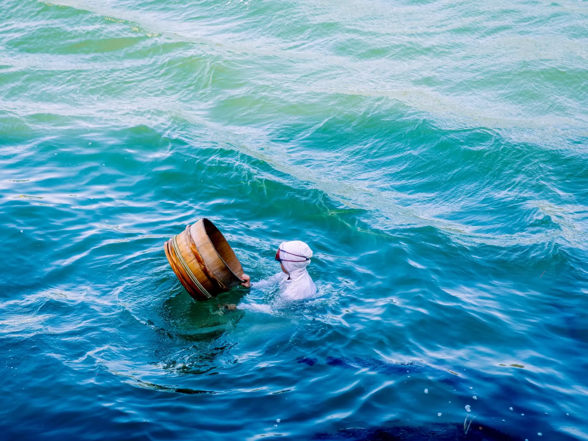 a women in a white garment with a hood and googles on her head swims in bright blue water beside a wooden bucket