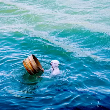 a women in a white garment with a hood and googles on her head swims in bright blue water beside a wooden bucket