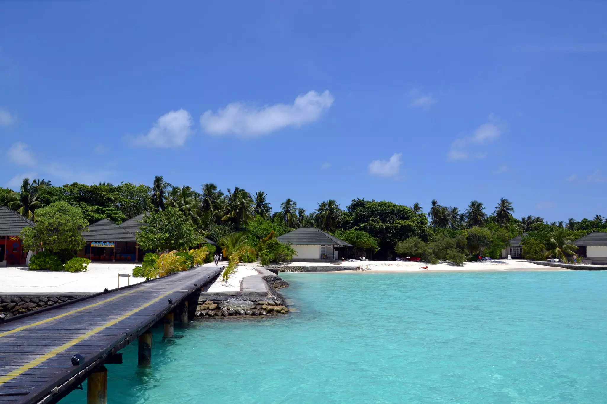 A dock over clear blue waters. Small building with sloped roofs dot the shore