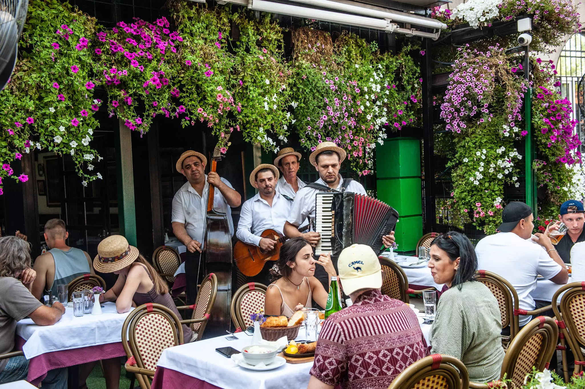 Folk musicians perform to diners sat at outdoors tables in a tavern covered in flowers.