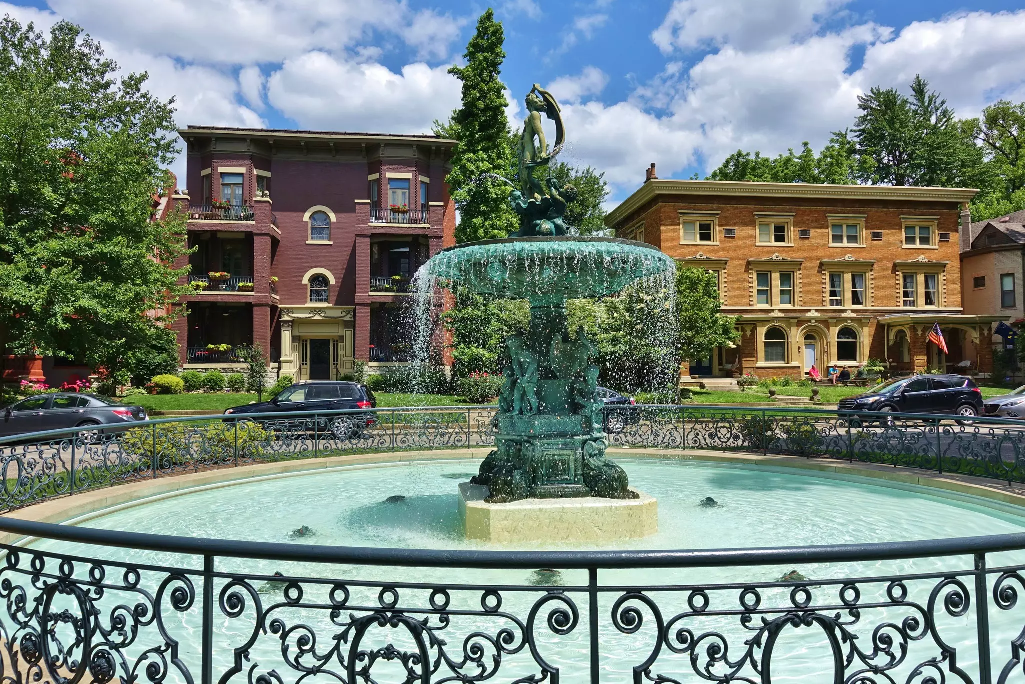 Wide shot of an ornate fountain in a round pool surrounded by a low metal fence with brick homes in the background.