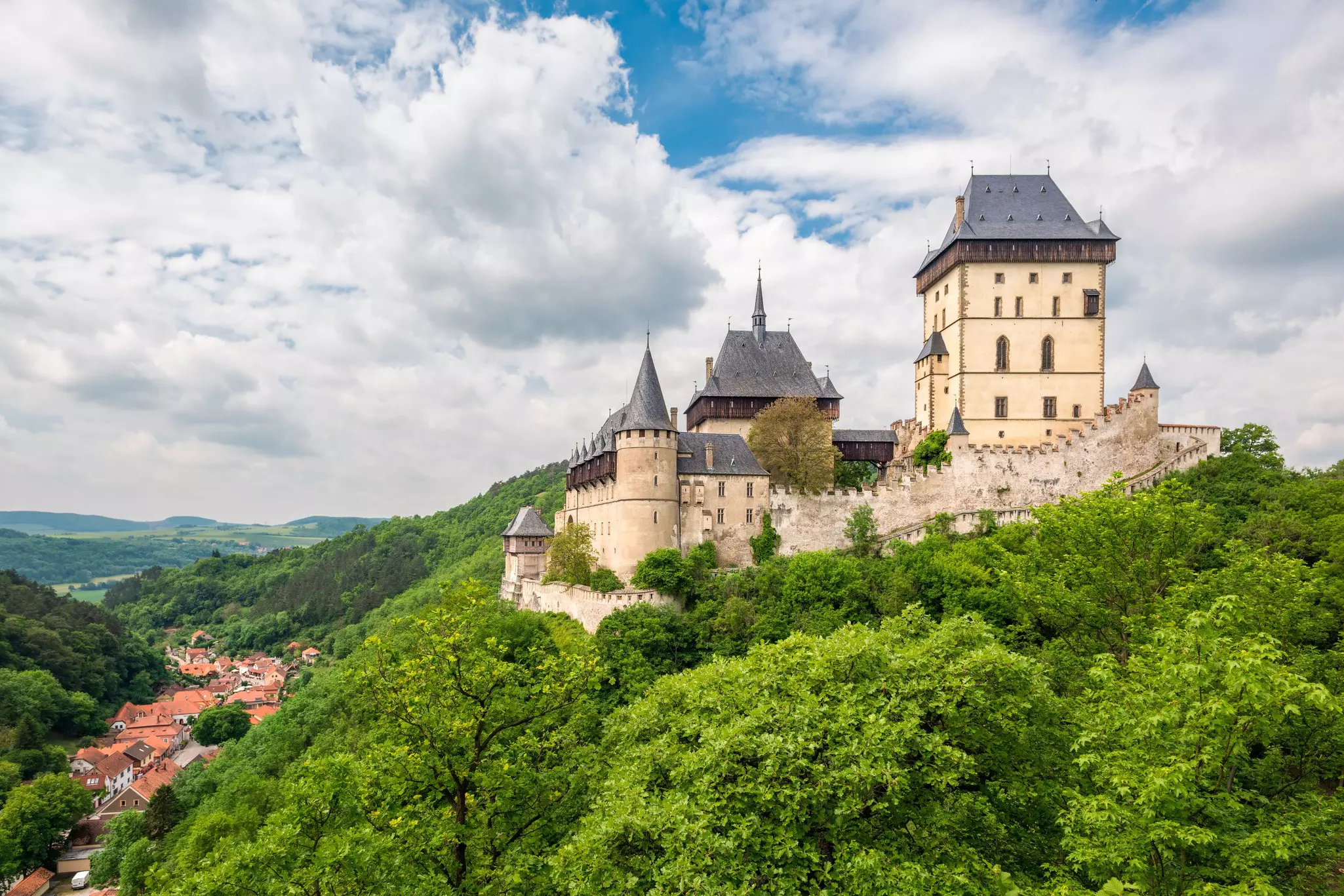 Towers of a stone castle are surrounded by a wall on a green hillside in Czechia.