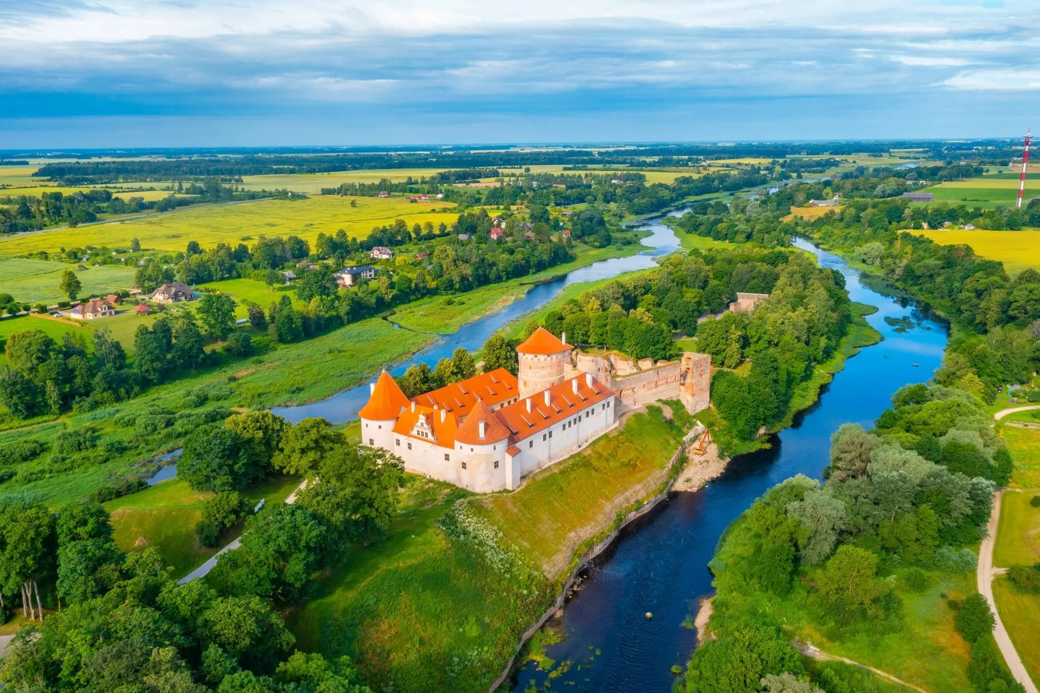 An aerial view of a castle with a red roof and turrets, on a spit of land between two small rivers in a landscape of green trees and fields.