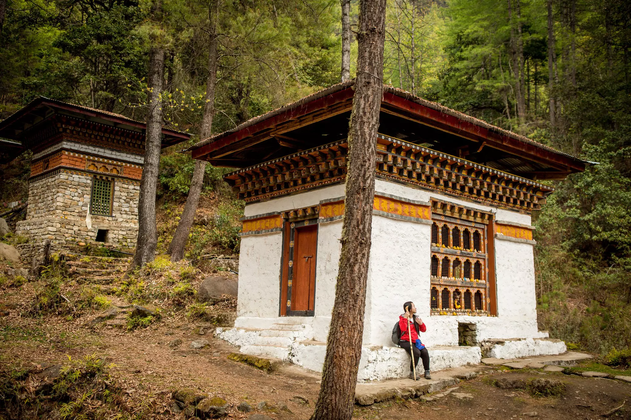A hiker rests, sitting on the ledge of a small white building in a wooded area.