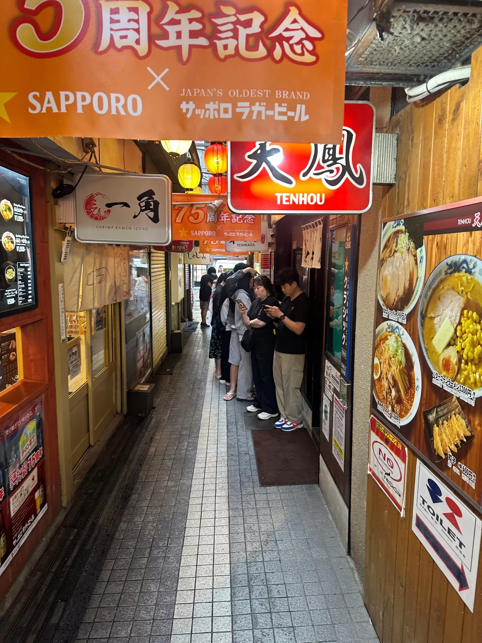 People form a line outside a noodle shop in a narrow lantern-lit alleyway: Ganso Ramen Yokocho. 