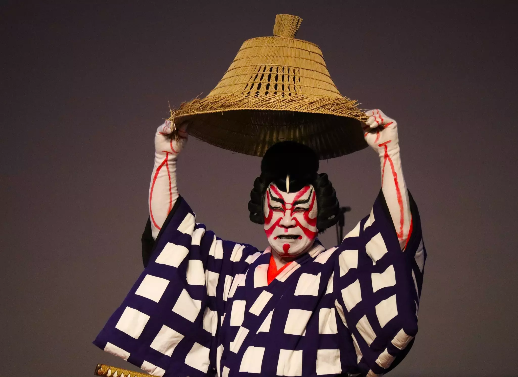 A performer in red face and white face paint and a checkered kimono holds a hat above his head.