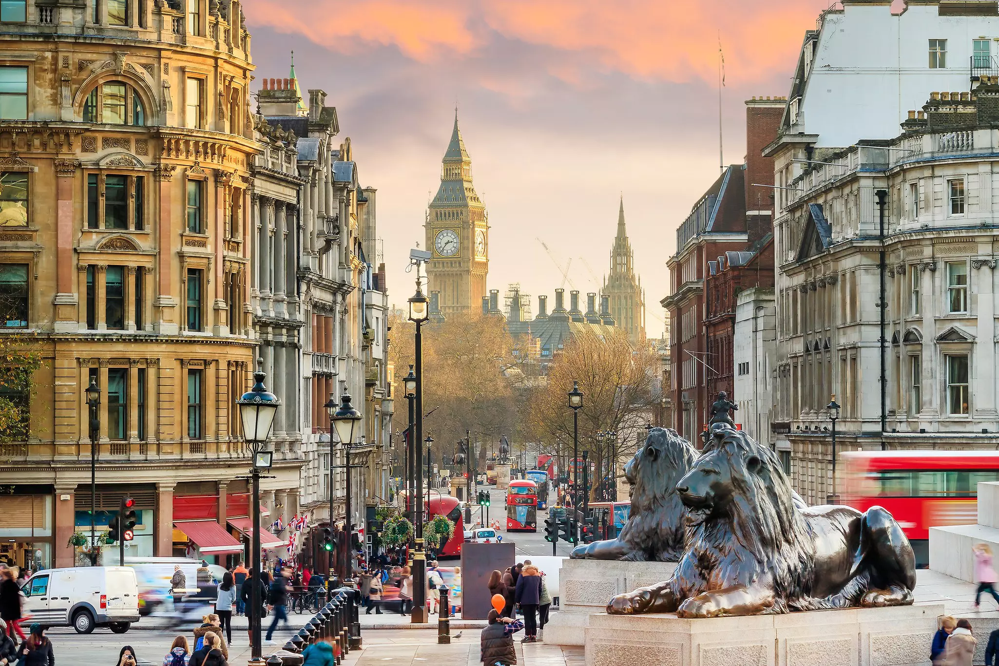 Large bronze lion sculptures in a public square. Double-decker red buses drive down the road and a tall Gothic clock tower stands in the distance