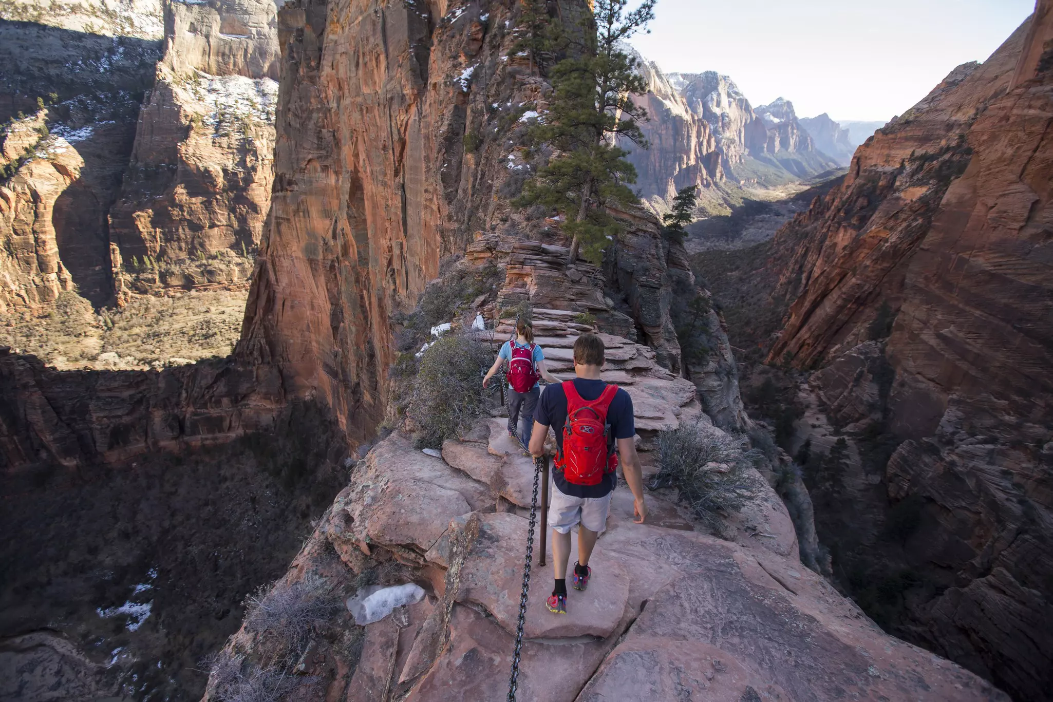 People are seen from behind hiking along a narrow, rocky ridge. Steep valleys are on either side of the path, and snow-dusted cliffs and mountains are visible in the distance.
