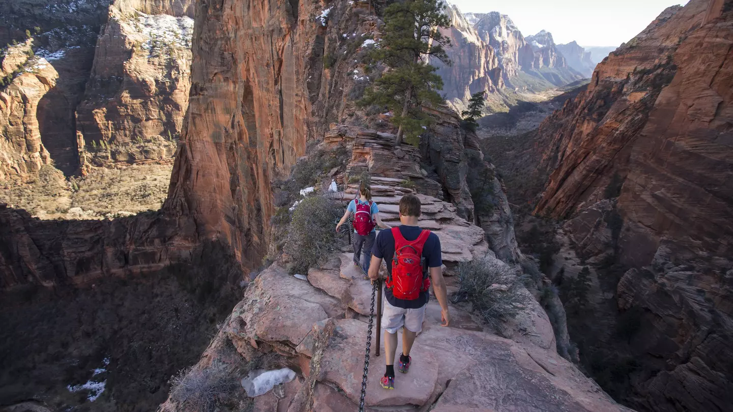 People are seen from behind hiking along a narrow, rocky ridge. Steep valleys are on either side of the path, and snow-dusted cliffs and mountains are visible in the distance.