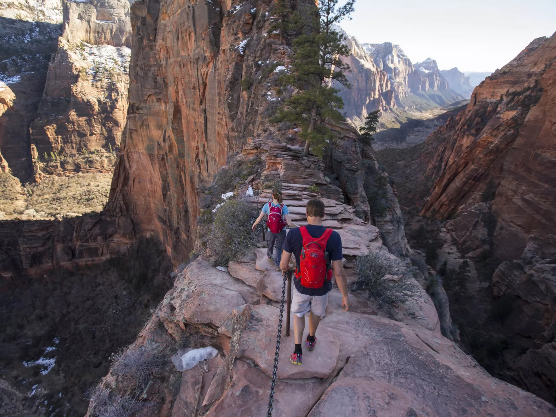 Hikers on a narrow, rocky ridge. Steep valleys are on either side of the path, and snow-dusted cliffs and mountains are in the distance.