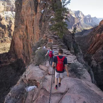 Hikers on a narrow, rocky ridge. Steep valleys are on either side of the path, and snow-dusted cliffs and mountains are in the distance.