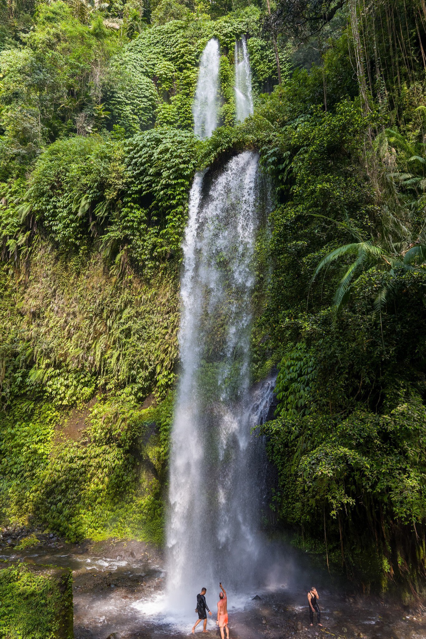 Two tourists standing at the base of towering Tiu Kelep waterfall in Senaru, Lombok, Indonesia.
