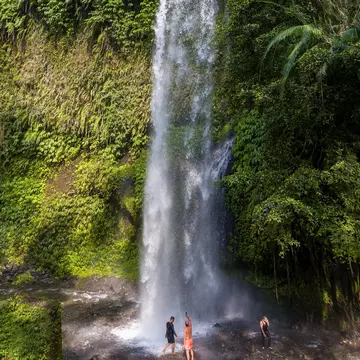Two tourists standing at the base of towering Tiu Kelep waterfall in Senaru, Lombok, Indonesia.