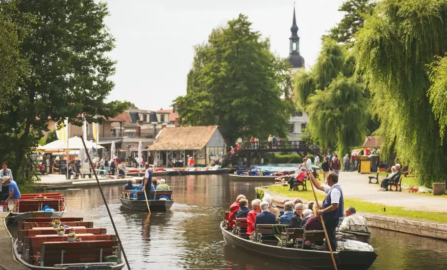 Passengers seated in a wooden punt poled along a tree-lined canal in the Spreewald, a church spire behind